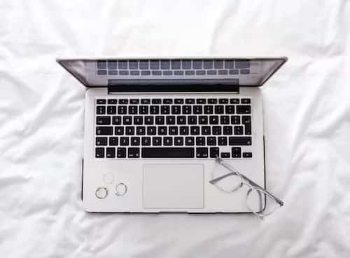 A closed laptop computer with a clean keyboard and glasses on a bed. The laptop is silver and has a black keyboard. 